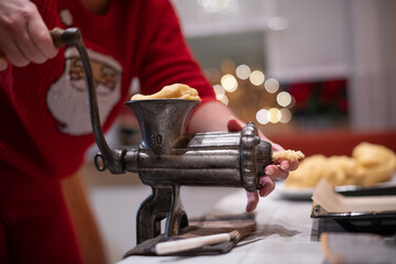 Nahaufnahme von Hände einer Frau, die mit einen alten Drehwolf, Spritzgebäck Plätzchen zu Weihnachten backt. Im Hintergrund das Bokeh von Weihnachtsbeleuchtung