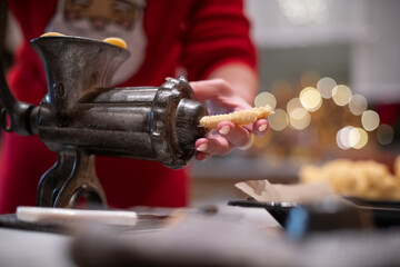 Nahaufnahme von Hände einer Frau, die mit einen alten Drehwolf, Spritzgebäck Plätzchen zu Weihnachten backt. Im Hintergrund das Bokeh von Weihnachtsbeleuchtung
