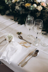 Wedding. Decor. Banquet. A table with a white tablecloth in the festive hall is decorated with a composition of flowers and greenery, there are candles, glasses, cutlery and name plates for guests