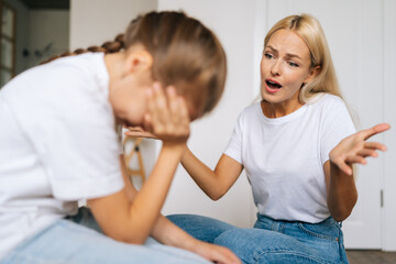 Fototapeta premium Close-up of sad depressed little girl crying covering face with palm while angry young mother scolding, raising voice, scream at stubborn difficult little child daughter for bad behavior at home.