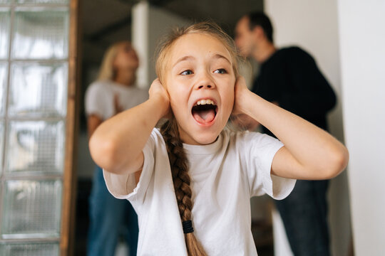 Close-up portrait of excited upset little daughter screaming looking away, covering ears while angry parents quarrelling and fighting on background at home. Concept of family problems, crisis.