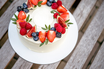 White circle tasty birthday cake decorated with fresh berries: strawberries, blueberries, raspberries and figs on round palte on wooden bench outdoors in summer day.