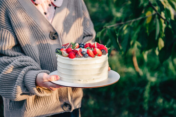 Unrecognizable caucasian girl wearing knitted cardigan holding white tasty birthday cake decorated with fresh berries outdoors in sunny day. Natural light and shadow