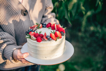 Unrecognizable caucasian girl wearing knitted cardigan holding white tasty birthday cake decorated with fresh berries outdoors in sunny day. Natural light and shadow