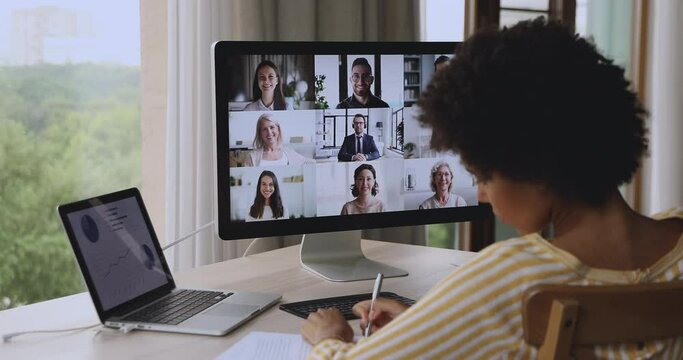 Group of diverse businesspeople involved in discussion, solve business remotely use videocall, pc screen view over African businesswoman shoulder sit at desk taking notes negotiates through video call