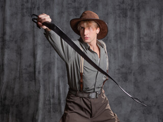 treasure hunter, a hero of a retro-style adventure. A young man in a hat and breeches with suspenders with a whip in his hands. Posing in the studio on a gray background - 554450386