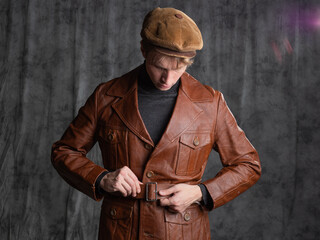 young man, a retro secret agent, a guy in a brown leather jacket and a flat cap with a gun in his hand. Posing in the studio on a gray background