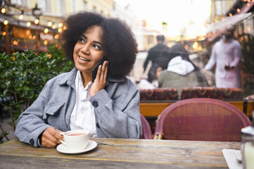 Fashion street style portrait of attractive young natural beauty African American woman with afro hair in tweed jacket speaks phone outdoors in sidewalk cafe. Happy lady with coffee cup in big city