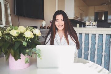 Young Beautiful Asian business woman working online with laptop computer placed at the table at home office. Smiling lady freelancer have remote work sitting in living room with white roses flowers