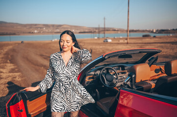 Happy smiling brunette woman driver in fashion dress posing with new red cabriolet car on beach...