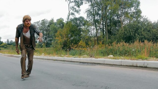 A drunk and dirty man walks along the road. An alcoholic and a homeless man covered in swamp strud walks down the street.