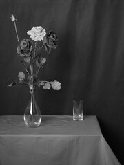 Künstliche Rosen in eine Vase neben einem leerem Glas auf bedeckten Tisch. Schwarzweis.