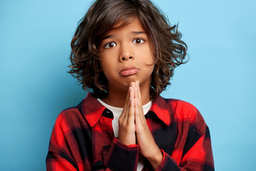 Close up shot of distressed teenager asks for help, keeps palms pressed together, purses lis, dressed in red checkered shirt, poses over blue background © South House Studio
