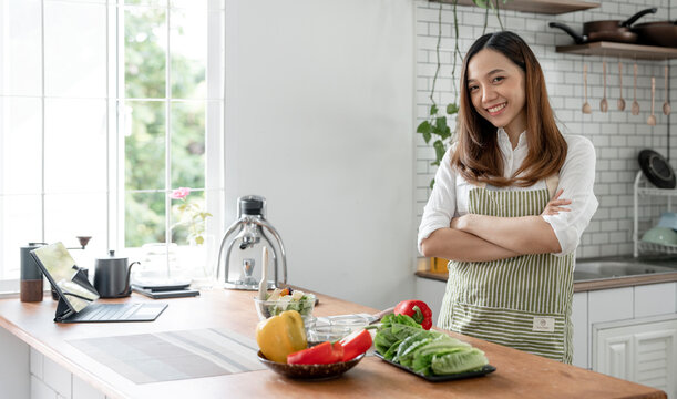 Portrait of young happy woman wearing appron standing with arms crossed in the kitchen room,  prepares cooking healthy food from fresh vegetables and fruits.