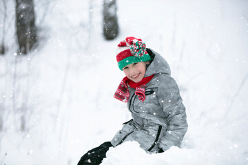 Child in winter for a walk. A little boy, in a red knitted hat, sits in a snowdrift.