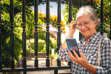 Beautiful smiling older woman standing in front of the gate of her house while using her mobile phone to write a message or watch social network content