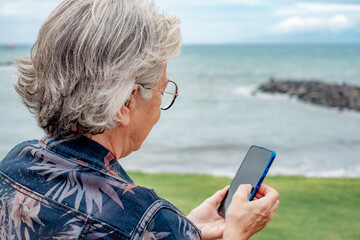 Rear view of senior woman sitting at the beach in a winter day using mobile phone. Trendy gray haired lady with eyeglasses relaxed in front to the sea enjoying tech and social on smartphone