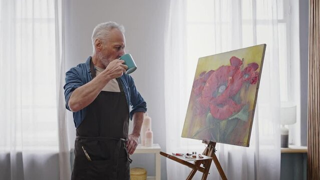 Retired senior artist drinks hot tea or coffee and looks at his author's painting with inspiration
