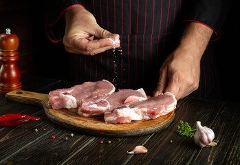 Professional chef salts raw sliced meat steaks on the cutting board of the restaurant kitchen before preparing dinner. Delicious grill idea
