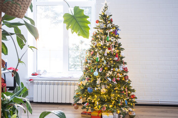 Christmas tree in the white interior of a house with large windows. Glowing fairy lights garlands interior decoration of the studio room. Potted plants in the home