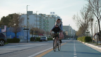 Mother rides bicycle with child in front in bike lane by sea shore. Parent riding bike outdoors during cold day
