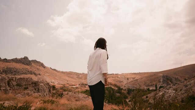 stehende Frau in weißem Hemd und schwarzer Hose genießt die Aussicht auf einem Hügel und betrachtet Gebirge und das Tal. Läuft dabei nach vorne.
