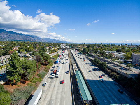 aerial shot of cars and trucks driving along the 210 Freeway surrounded by lush green trees and plants, apartments and office buildings with mountains, blue sky and clouds in Pasadena California