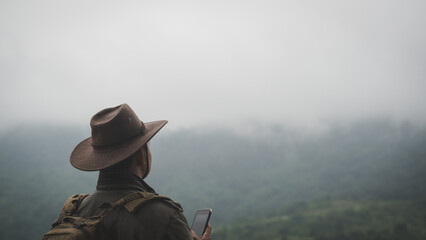 Freedom traveler man in hat carrying a backpack stands at the top of a mountain and using a smartphone on a foggy day.Adventure travel and success concept