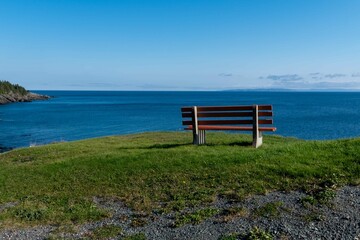 An empty orange wooden bench with metal posts on long lush green grass with small yellow flowers. The seat has a deep blue ocean in the background with a long landmass and blue sky in the background. 