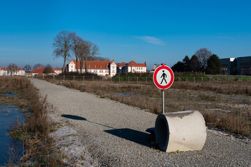 A prohibitory sign for pedestrians and a concrete ring on a lonely path leading to a construction site. In the background is a building under a blue sky.