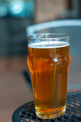 A clear pint drinking glass filled with cold froth from a lager ale. The Irish red ale pint sits on the edge of a metal patio table at a microbrewery. There are tables and chairs in the background.