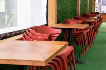 Red plastic patio chairs and square wooden tables are stacked against a wall in an outdoor restaurant. There's a green golf carpet on the decks floor. The piled retro terrace bistro seating is empty.