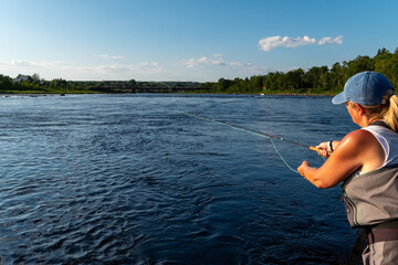A middle age female stands in a large salmon river casting a fishing rod holding line in her left hand. The blonde long haired lady is wearing a blue hat, white t-shirt, and waders fly-fishing.