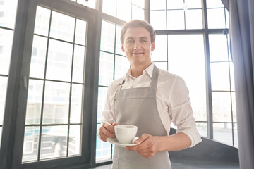 A waiter in a gray apron holds out a cup of coffee. A barista serves a cup of hot coffee in a cafe,...