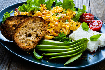 Breakfast - scrambled eggs with bacon, cottage cheese, avocado and vegetables on wooden table
