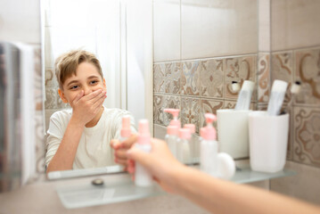 Obraz na płótnie Canvas Teenager boy looking at the mirror, smiling and touching his face and holding cosmetic bottle in bathroom. Beautiful healthy child at home doing his morning skincare routine and having fun.