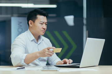 Worried young Asian man sitting at the desk in the office. He holds a credit card in his hand, looks at the laptop, checks the account, pays for purchases.