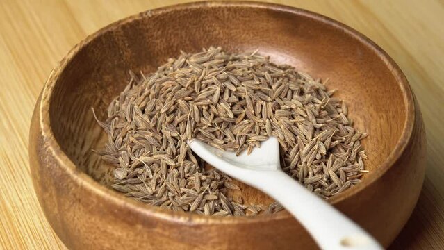 Dried cumin spice seeds in wooden bowl with spoon rotating on wooden board. Dry seasoning in spoon. Spices and herbs for cooking zira