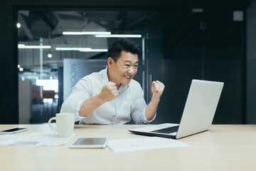 A young Asian man sits in the office at the table, during working hours he watches a football match, sports competitions,makes bets, cheers and rejoices on his laptop.