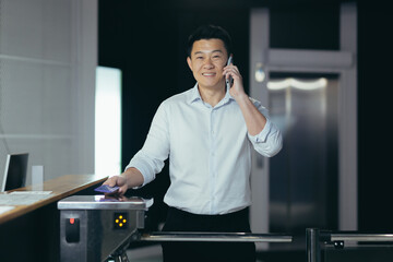 A young Asian man, the director,businessman, enters the office through an electronic turnstile, an automatic security system. Encloses his electronic access card. Talking on the phone, smiling.