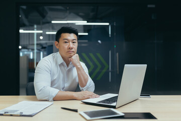 Fototapeta na wymiar Portrait of a young handsome successful Asian man, businessman, company founder, office worker sitting at a desk in the office, looking confidently into the camera, holding his head with his hand.