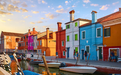 Fototapeta na wymiar Burano island in Venice, Italy. Motley coloured home coast with boats floating on Channel, blue sky and evening sunset