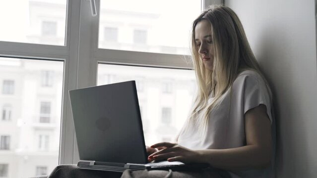 A young woman is sitting on a windowsill, messaging on a computer, working. Distant work.