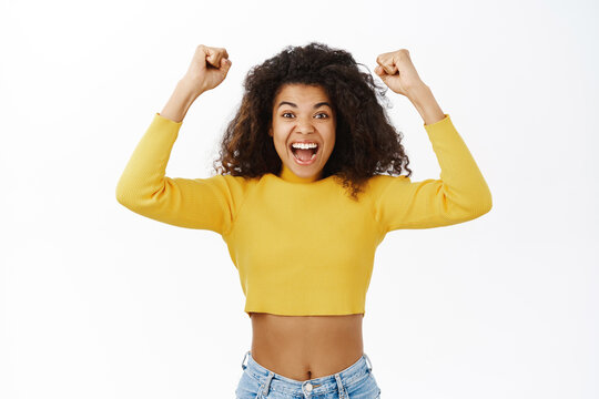 Enthusiastic woman celebrating, winning and triumphing, achieve goal, smilng and looking in disbelief, standing against white background
