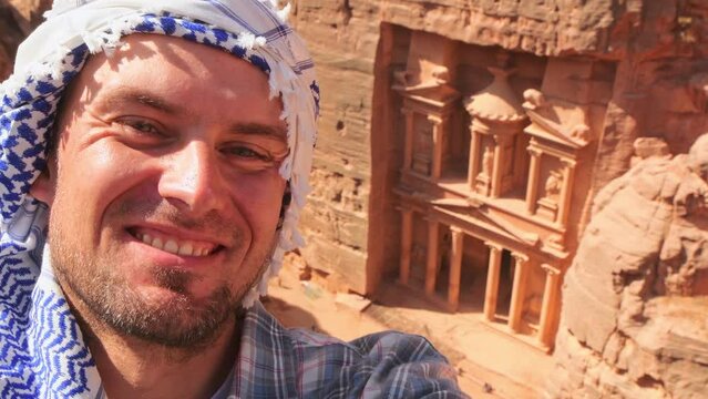 Caucasian male tourist stand on viewpoint in Petra ancient city over Treasury or Al-khazneh take smartphone photo smile. Jordan, one of seven wonders. UNESCO World Heritage site.