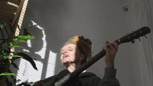 woman with dreadlocks on head singing song and playing acoustic guitar in sunny cottage bedroom, simple living and hobby relaxing