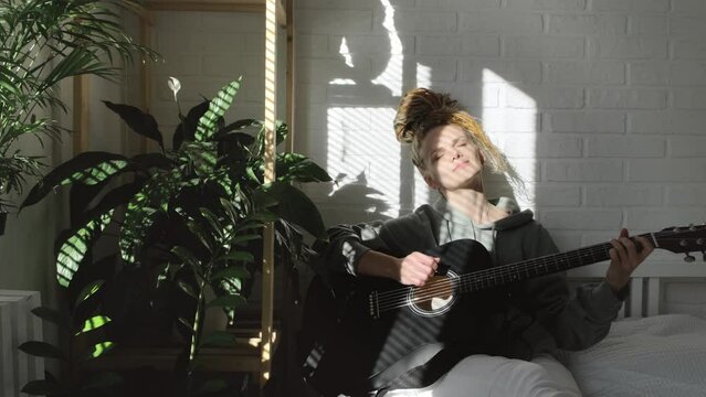woman with dreadlocks on head singing song and playing acoustic guitar in sunny cottage bedroom, simple living and hobby relaxing