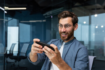 Mature businessman with beard is playing game on smartphone, man is having fun during break at workplace, businessman inside modern office.