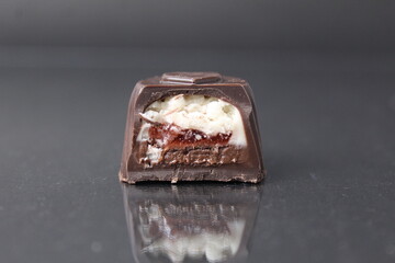 a chocolate candy case creamy slice in a section on a black background and a whole candy next to each other. White milk chocolate filling with coconut praline berry cherry jelly. Chocolate Sweets Day