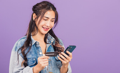 Portrait of Asian beautiful young woman confident wear denim jeans shirt smiling using mobile phone holding credit card bank, Happy female hold smartphone, studio shot isolated on purple background